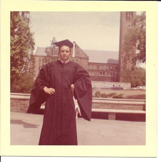 A colour snapshot with a white border: a young Roger Fogelman in a black cap and gown, standing on a stone terrace on the Cornell campus, with a stone building and the campus bell tower behind him.